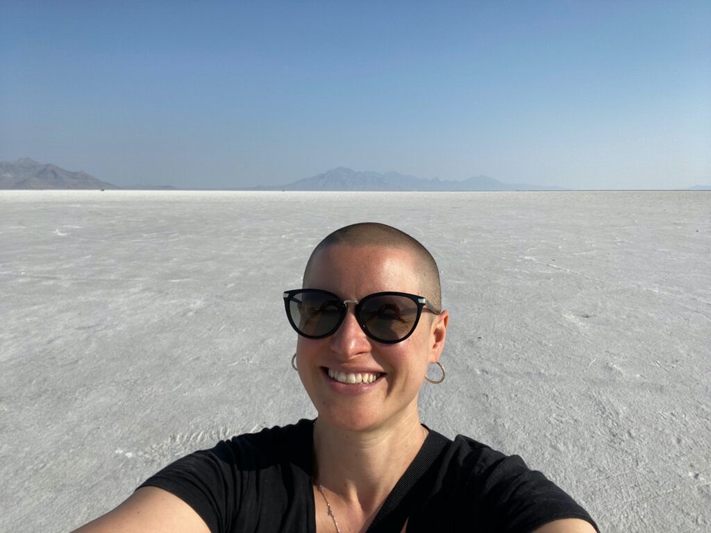 Sarah Dopp, a white woman with a shaved head, is wearing sunglasses, hoop earrings, and a black t-shirt and smiling for a selfie. There is a large empty stretch of salt flats in the background.
