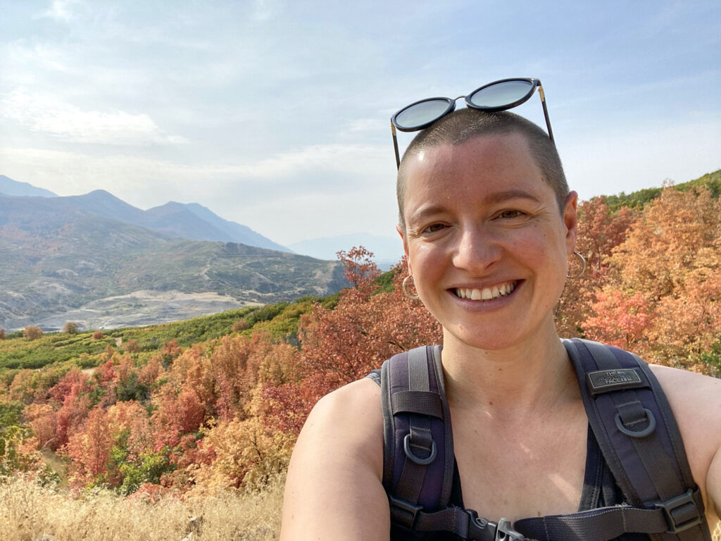 Sarah Dopp is standing on the top of a hill on a hike with autumn leaves in the background. She is wearing a backpack and has sunglasses on top of her head. She is smiling.