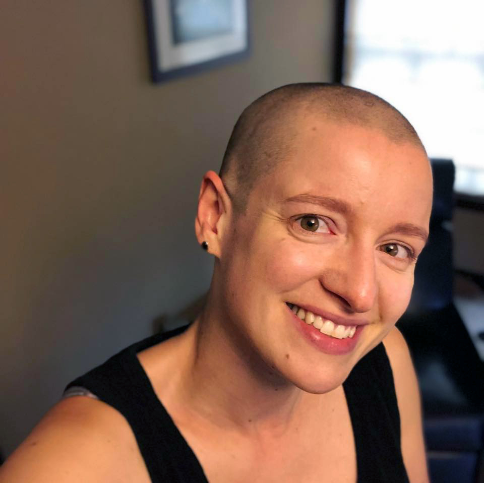 A white woman with a shaved head wearing a black tank top, smiling