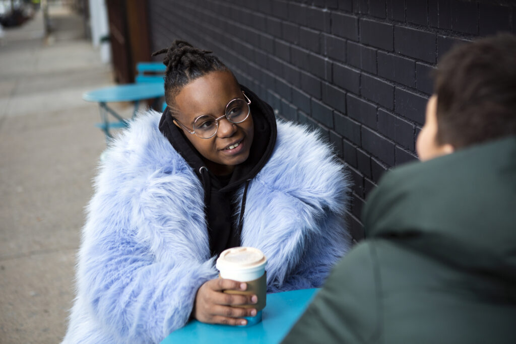 A transmasculine person with a furry blue coat drinking coffee with a friend